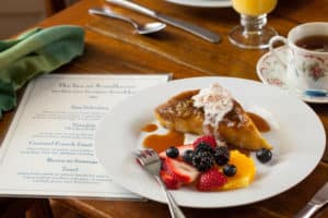 Wooden breakfast table set with a white plate of French toast casserole topped with cream and an assortment of berries, plus a white menu, cup of tea, and glass of orange juice.